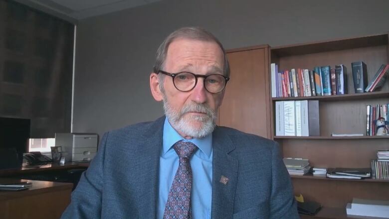 A man with a grey beard wearing a suit and glasses speaks in an office setting.  