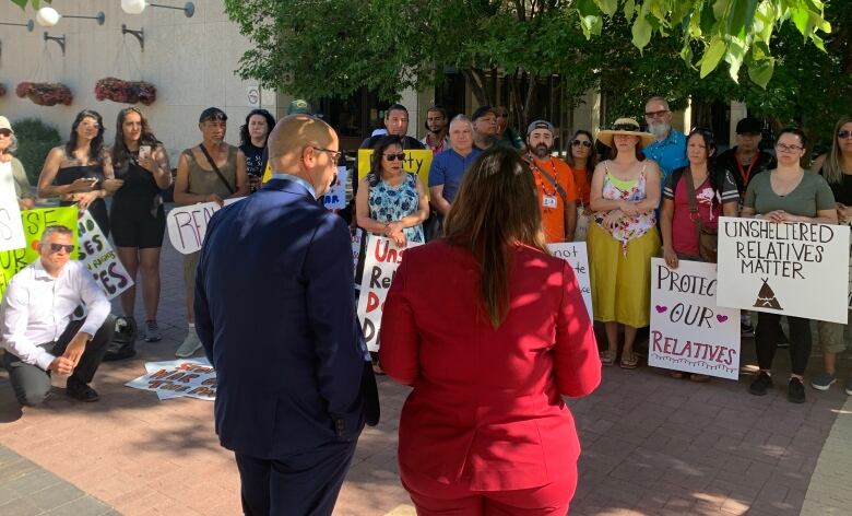 A man in a dark blue suit and a woman in a red suit face a group of people in a courtyard holding signs with messages like 'Protect Our Relatives.'