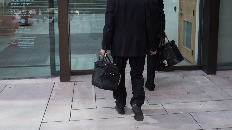 Men wearing black robes and black shoes carry black briefcases into a glass courthouse.