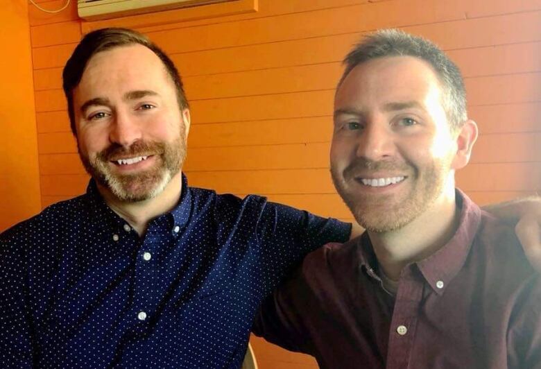 Two men sit facing the camera, smiling. They are sitting indoors, with their arms around each other's shoulders, against the backdrop of an orange wall. 