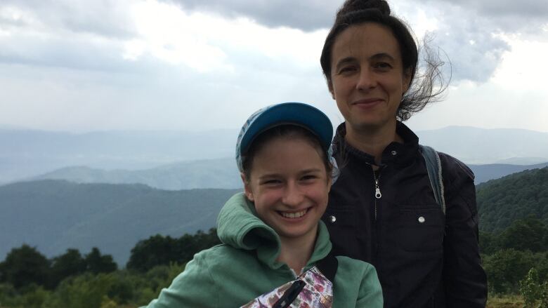 A girl and her mother smile for the camera with hills behind them in the distance.