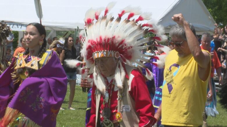 Three people performing during grand entry of Sitansisk Wolastoqiyik powwow near Fredericton. Two people are wearing regalia. On the far left a women can be seen in braids and in purple regalia with yellow, blue and gold beading. In the middle, an elderly man is wearing red regalia with a red headdress. On the right, someone can be seen wearing a yellow tank top and sunglasses while raising a fist.    