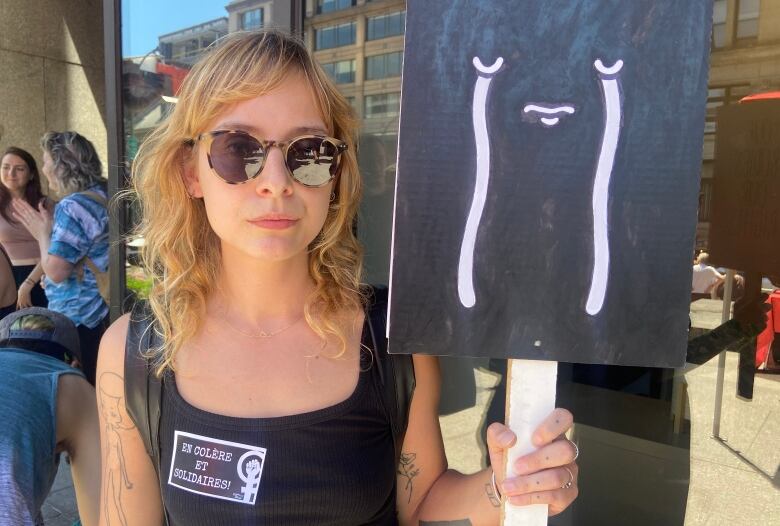 A person with blond hair and sunglasses holds up a black and white sign with a crying emoji. 