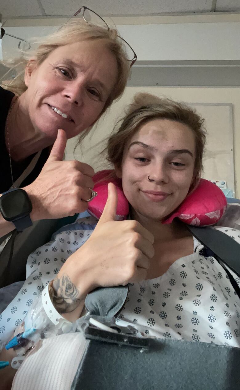 Two women, one in a hospital gown, smile for the camera and give the thumbs up sign.