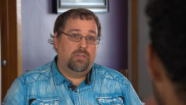 A middle-aged man wearing glasses and a blue shirt speaks with a reporter.