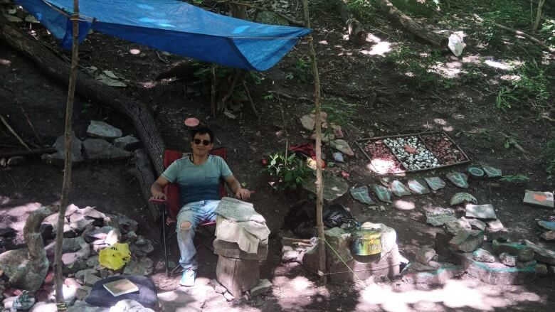 A man sits under a tarp along a path in the forest, surrounded by painted rocks on the ground among the trees. 