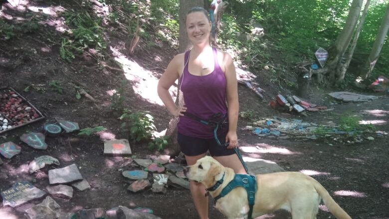 A woman stands on a forested path with her hand on her hip, her other hand holding a leash the leads to her dog.