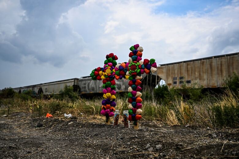 Two colourful crosses and a pair of candles stand on the ground in front of a line of train cars parked on the tracks. 