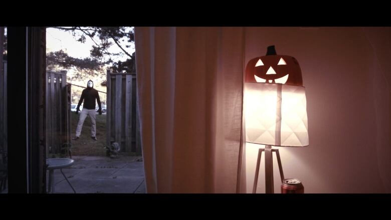 A hooded, masked figure looms in front of an open door, a jack o lantern and lamp are in the foreground.