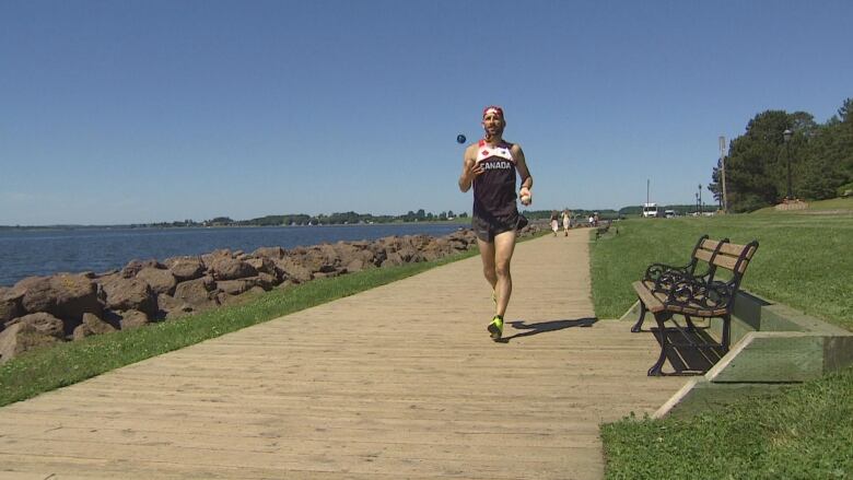 Man in baseball hat, tank top, and shorts running down an ocean-side walking trail while juggling.