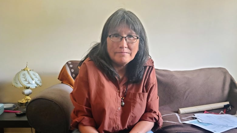 A woman with grey hair sits on a couch. A pair of moccasins and a high school diploma rest on the coffee table in front of her.