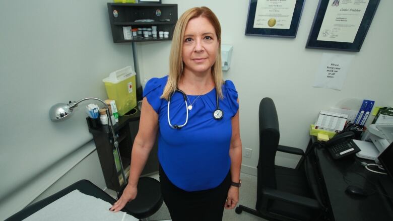 A woman wearing a stethoscope stands in a medical examination room.