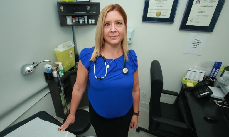 A woman wearing a stethoscope stands in a medical examination room.