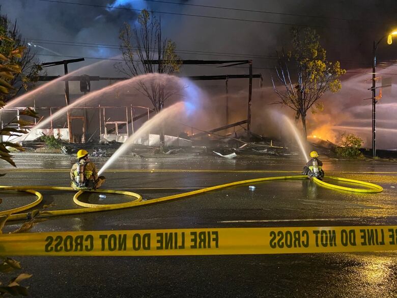 Two firefighters lie on the ground, spraying hoses at a gutted building behind a yellow line that reads 'Fire line do not cross'.