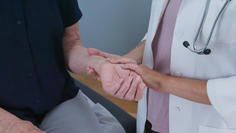 A closeup photo shows a doctor in a lab coat, with a stethoscope around her neck, holding the arm of a patient and taking her pulse.