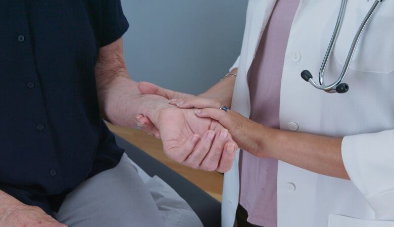 A closeup photo shows a doctor in a lab coat, with a stethoscope around her neck, holding the arm of a patient and taking her pulse.