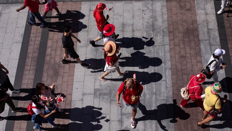 An aerial shot of people crossing a sidewalk. Most of them are wearing red in celebration of Canada Day. A person on the right has a hat with a Maple Leaf on it, and a man in the centre has a tiny Canada flag sticking out of his backpack.