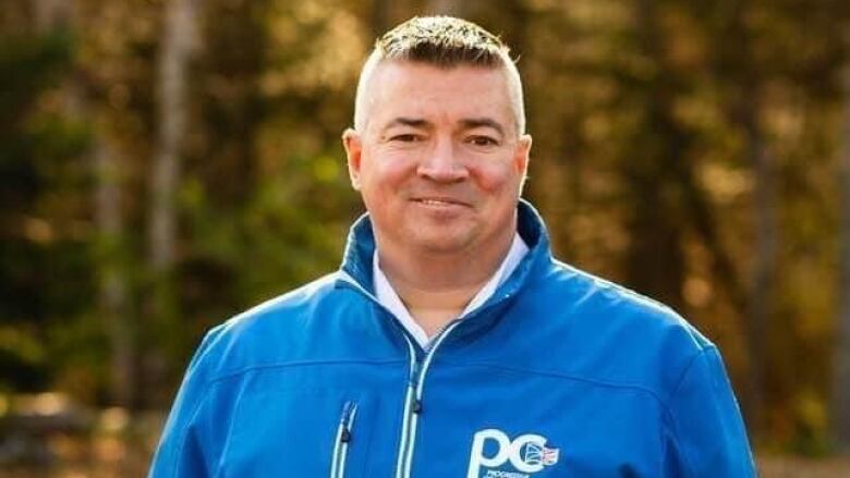 Man in blue coat with trees behind him.