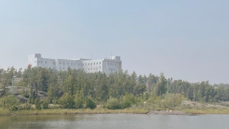 A hospital is seen from across a river through hazy smoke.