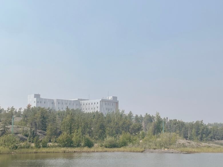 A hospital is seen from across a river through hazy smoke.