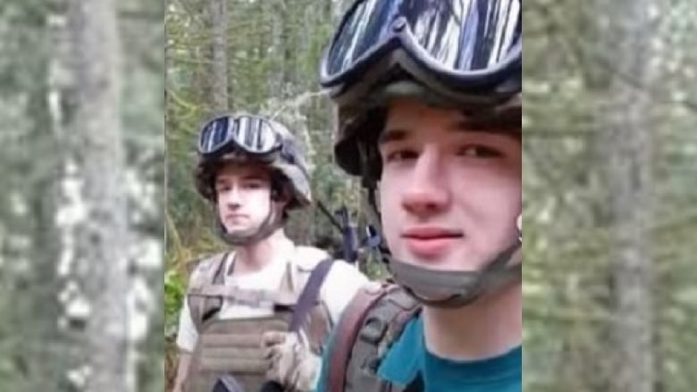 Two young men stand in a forested area wearing large helmets with chin straps.