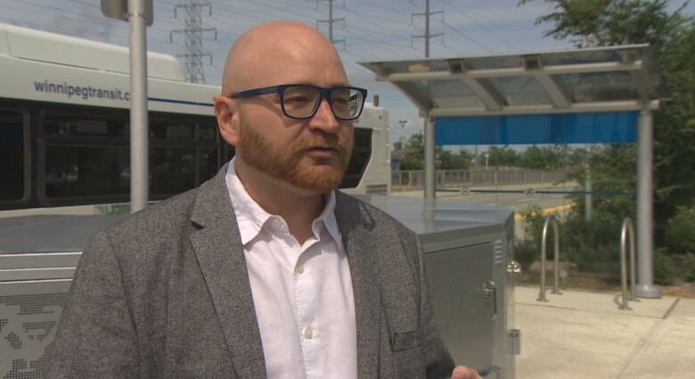 A bald man with a beard and dark-framed glasses stands in front of a bus stop.