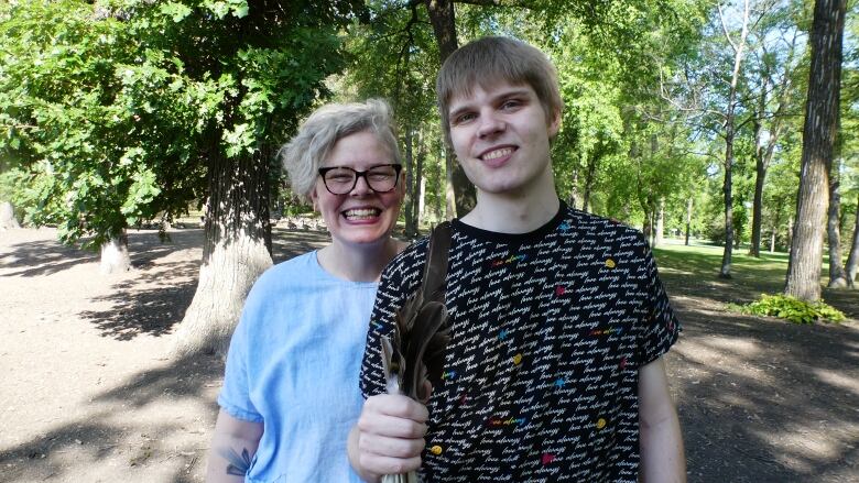 Kalyn Falk and her son Noah are shown standing side by side in a park with trees in the background. Noah is holding up a handful of feathers towards the camera and they are both smiling.