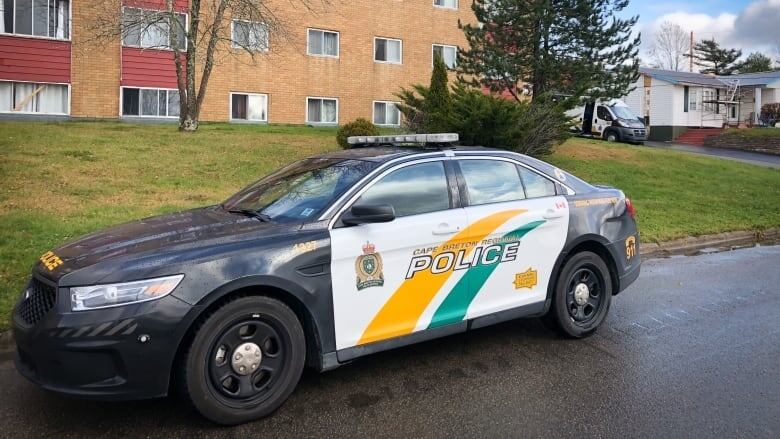 A marked police car is parked on a road by a lawn and a brick apartment building. 