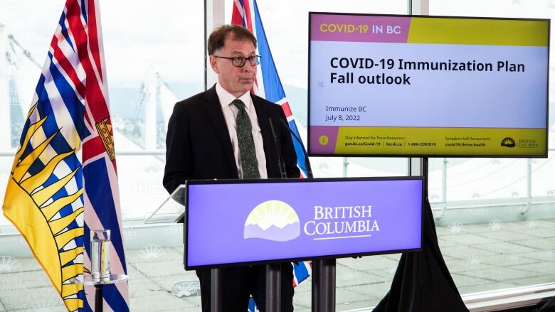 Adrian Dix stands in front of a podium with the logo and words 'British Columbia' on it. He is a white man with brown scattered hair and square glasses, wearing a black suit with a speckled tie. Behind him, two British Columbia flags are hung, in addition to a TV screen with the words 'COVID-19 Immunization Plan Fall outlook'.