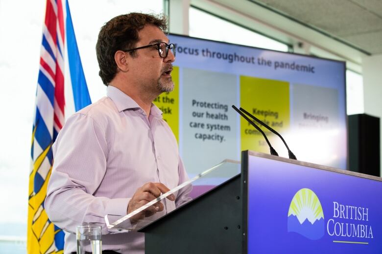 Dr. Martin Lavoie stands in front of a podium with the logo and words 'British Columbia' on it. He's a white man with a salt-and-pepper French beard and square glasses, wearing a pink full-sleeved shirt. Behind him, a B.C. flag and a TV screen with government focuses are listed. Focuses include 'protecting our health care system capacity'.