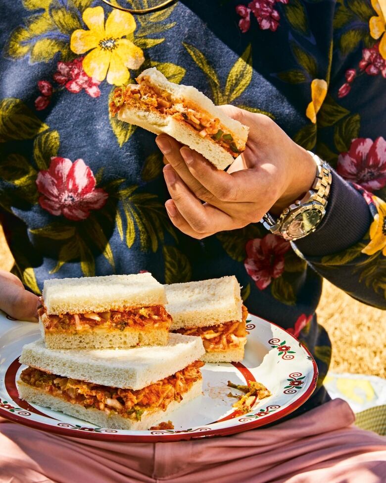 closeup on the torso of a man wearing a floral sweater and pink pants sitting outside at a picnic, holding a kimchi sandwich