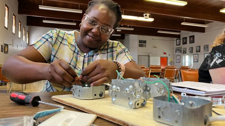 Mpokeleng Makwati works with electrical tools, with a screwdriver nearby. She's wearing safety glasses and smiling at the camera. 