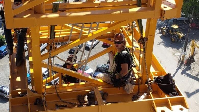 A man wearing a hard hat and helmet is shown sitting in a structure composed of yellow girders that are part of a crane assembly.