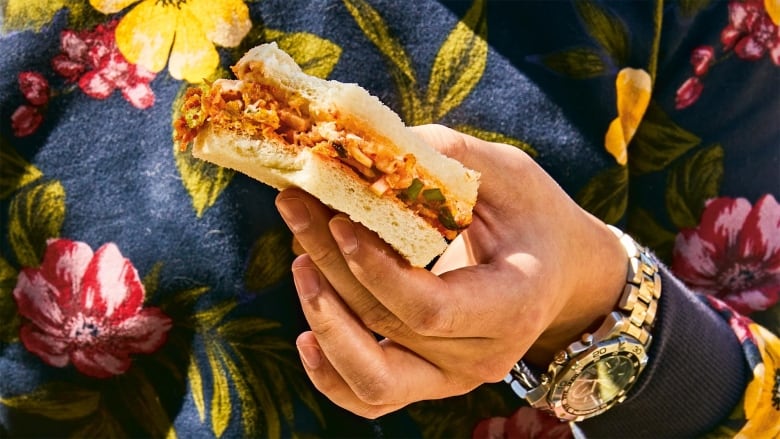 closeup of a hand holding a kimchi sandwich. the person is wearing a floral sweater and a watch.