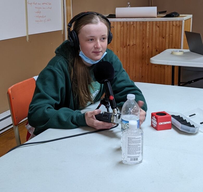 A teenager wearing headphones sits at a table holding a recorder, with a microphone in front of her.