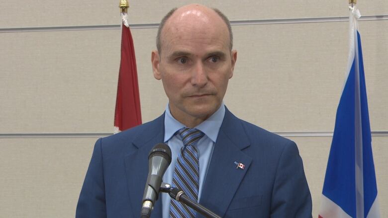 A man wearing a blue suit speaks with reporters in St. John's. He stands in front of two flags, one of Canada and one of Newfoundland and Labrador.