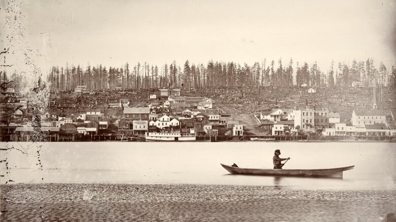 An archival picture of the city of New Westminster, in sepia tone. It shows a person boating along a shallow river, with houses built on the hill in the background.