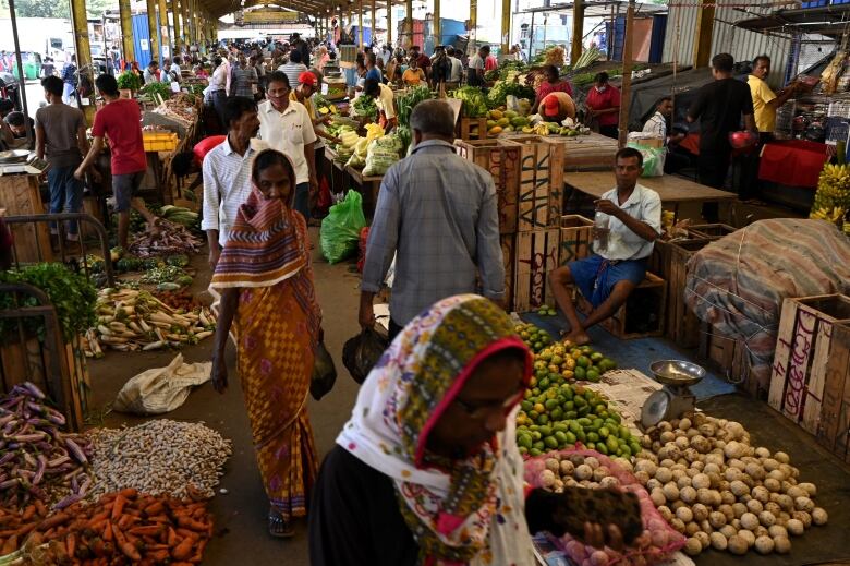 People walking through a vegetable market with piles of fruits in vegetables on the ground and on tables. 