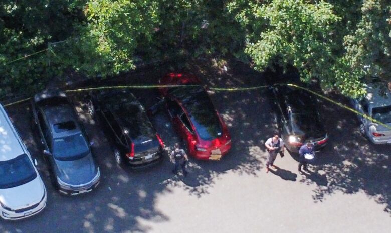 An aerial photograph of a number of cars parked at a parking lot, encircled by yellow crime tape. A few police officers are seen near a red sedan.