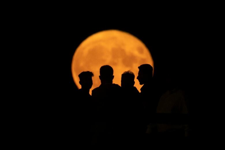 A full moon known as a buck moon rises behind a pier in White Rock, British Columbia on Wednesday, July 13, 2022. See large orange moon with silhouette of four people in front.