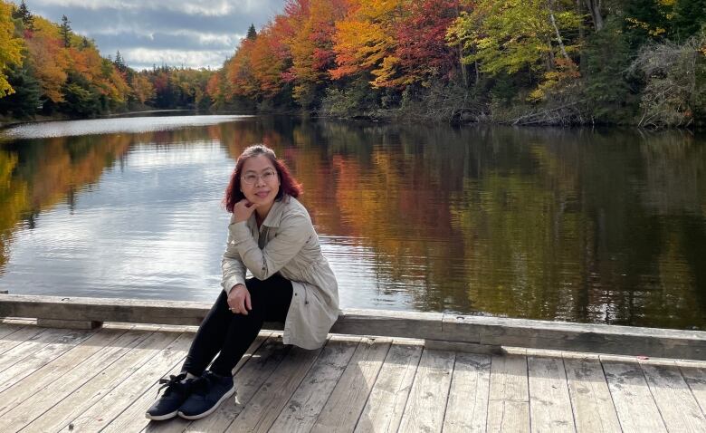 Vietnamese woman with glasses sits on dock over water. Colourful autumn colours surround the water.