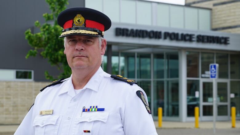 A portrait of a man, wearing a police uniform including a hat, in front of a police station. 