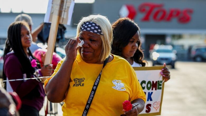 A woman wipes tears from her eyes outside a supermarket. Another woman behind her holds a sign that says 