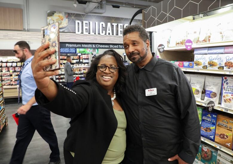 A woman holds up an smartphone to take photo with a tall, bearded man inside a supermarket.