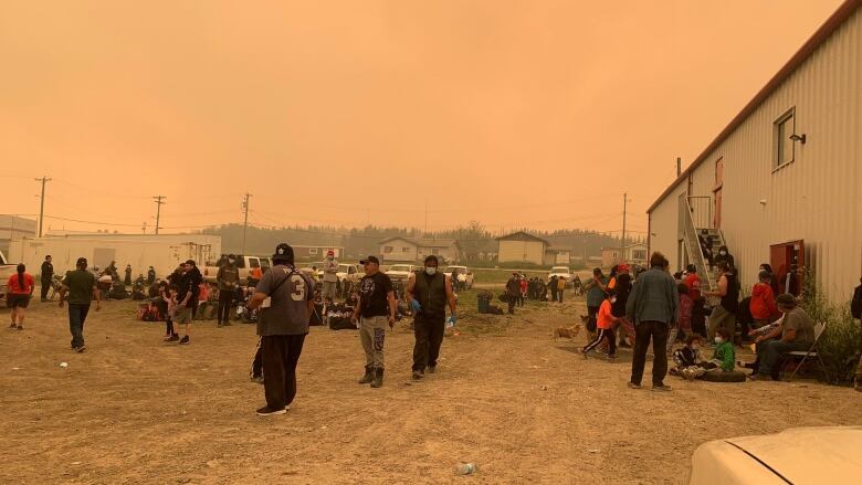 Dozens of people standing outside on a gravel yard with buildings in the background. The sky above is orange and filled with smoke.