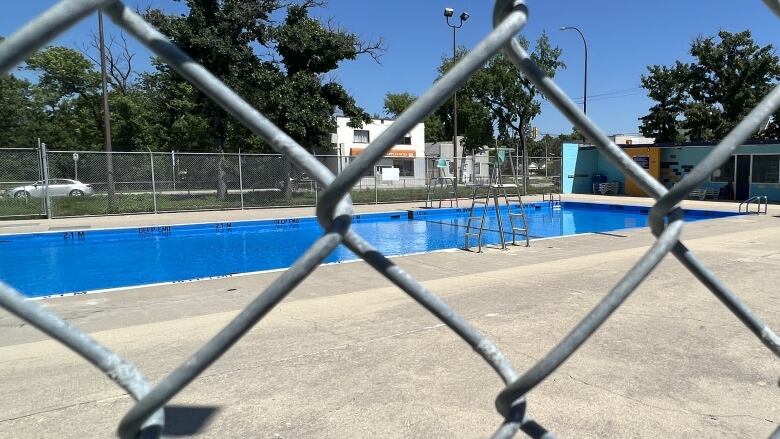 An empty outdoor swimming pool is shown behind a chain link fence on a sunny day.