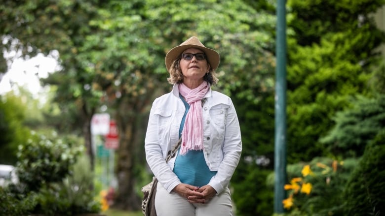 Susan MacRae, a blonde woman wearing a wide-brimmed hat, is pictured in the West End in Vancouver on Monday, July 18, 2022. MacRae stands in front of a blurred background of trees and flowers, and is dressed in light pants, a white denim jacket, turquoise shirt and pink scarf.