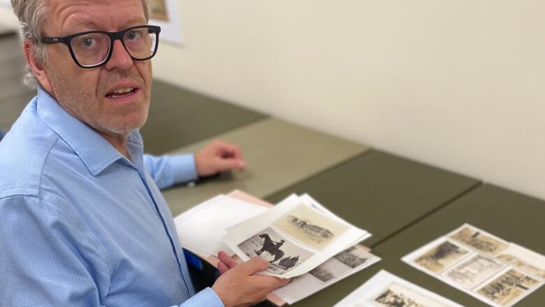 A grey-haired man in shirt and jeans stands by an open filing cabinet, examining archival photos.