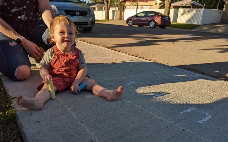 A small child holds coloured chalk and is sat on the sidewalk