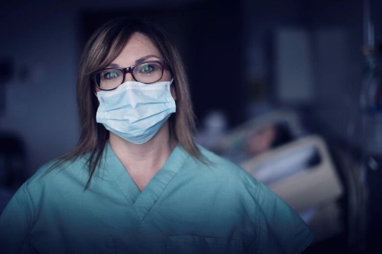 Nurse Niki Parsons is wearing a mask in the foreground with a patient in a bed in the background. 